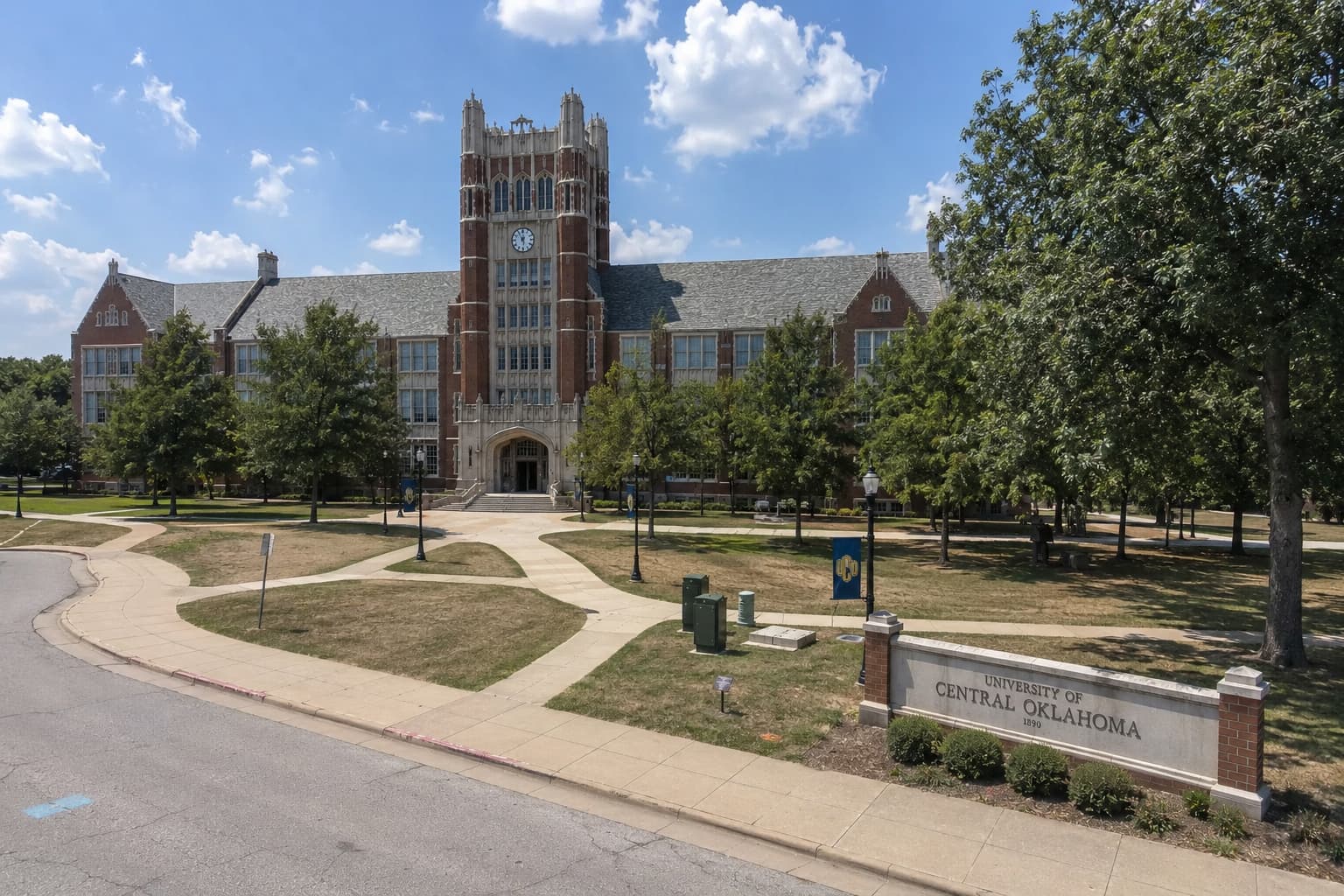 オクラホマ州エドモンドにある University of Central Oklahoma（UCO）の Old North 周辺をイメージした夏季キャンパス風景。赤レンガと石灰岩装飾の歴史的校舎、乾いた芝生のクアッド、曲線状の歩道、UCO サインモニュメントが広がる大学景観